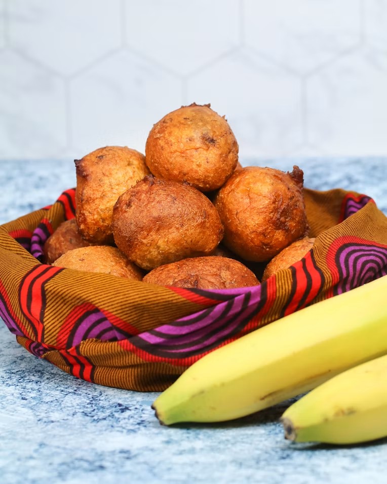 A basket of golden fried puff puff dusted with sugar, lined with African wax-print cloth, with two bananas beside it